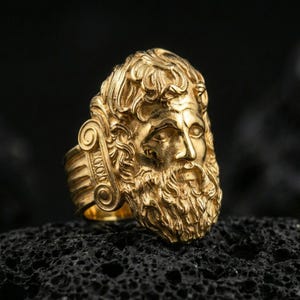 Gold plated Zeus signet ring with high-relief Greek god head and intricate hair detailing, premium studio macro on black volcanic rock surface