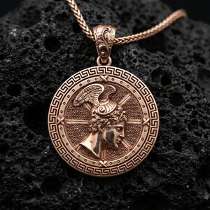 Rose gold plated Perseus coin pendant necklace on textured volcanic rock, centered studio-style close-up showcasing soft rosy glow and intricate Greek mythology portrait detailing.