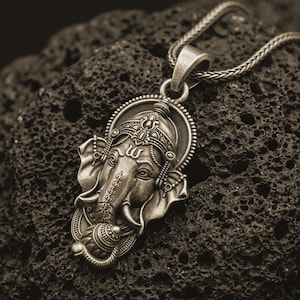 Bronze Ganesha pendant necklace on textured volcanic rock, close-up studio shot highlighting the elephant god face, ornate crown, tusks, and antique silver contrast.