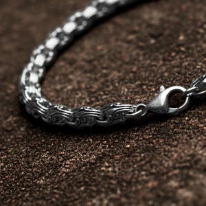 Può includere: Primo piano di un braccialetto color argento con un design ornato e strutturato. Il braccialetto presenta un motivo dettagliato e una chiusura sicura. Il gioiello è esposto su una superficie marrone strutturata, evidenziando gli intricati dettagli della lavorazione del metallo.