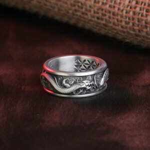 May include: A silver ring with a detailed dragon design wrapping around the band. The ring has a textured surface and intricate carvings, showcasing the dragon's scales and features. The top of the ring features a geometric pattern.