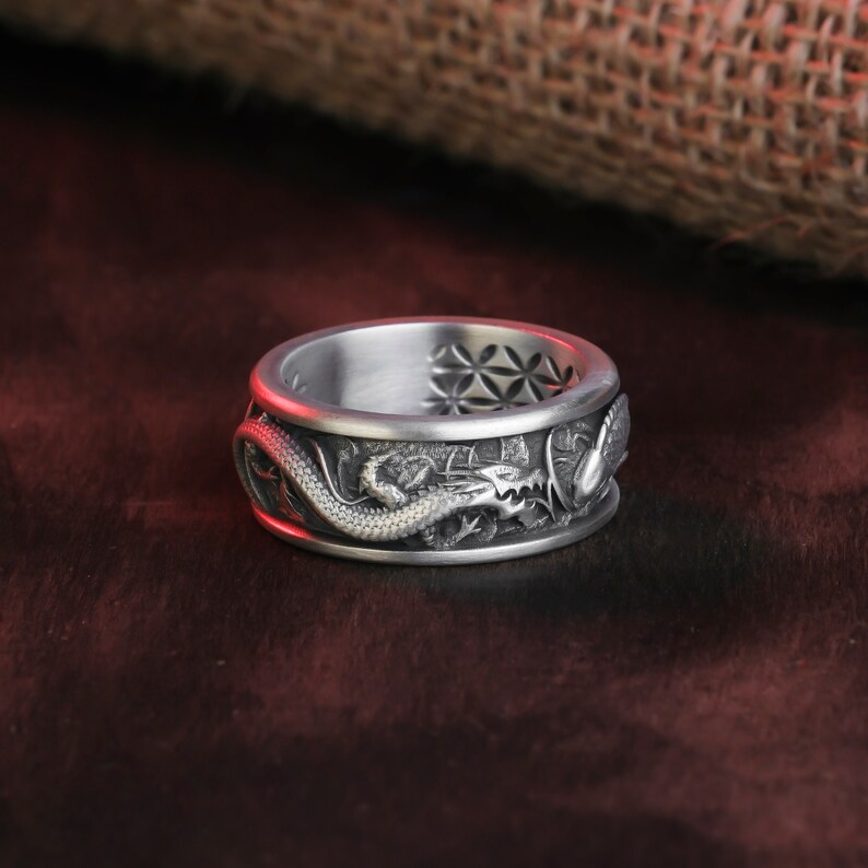May include: A silver ring with a detailed dragon design wrapping around the band. The ring has a textured surface and intricate carvings, showcasing the dragon's scales and features. The top of the ring features a geometric pattern.