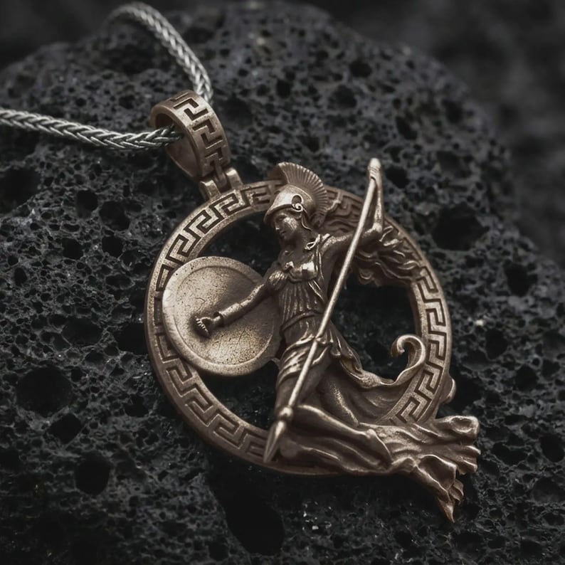 Bronze Athena warrior pendant necklace on textured volcanic rock, centered close-up highlighting the Greek key frame, helmeted goddess figure, spear, and deep antique shadowed contrast.
