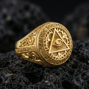 Gold plated all-seeing eye pyramid ring with detailed scrollwork band and Latin text around the face, close-up on volcanic rock