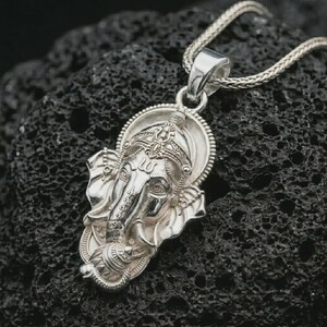 Polished silver Ganesha pendant necklace on textured volcanic rock, centered macro jewelry photo with detailed Hindu deity relief and bright silver highlights.