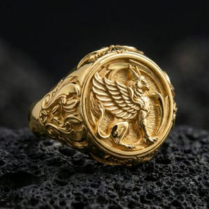 Gold plated griffon signet ring with high-relief mythical griffin motif and ornate border, premium studio macro on black volcanic rock surface