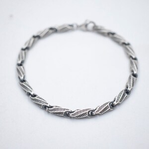 May include: A silver bracelet with a chain link design featuring a repeating Greek key pattern.