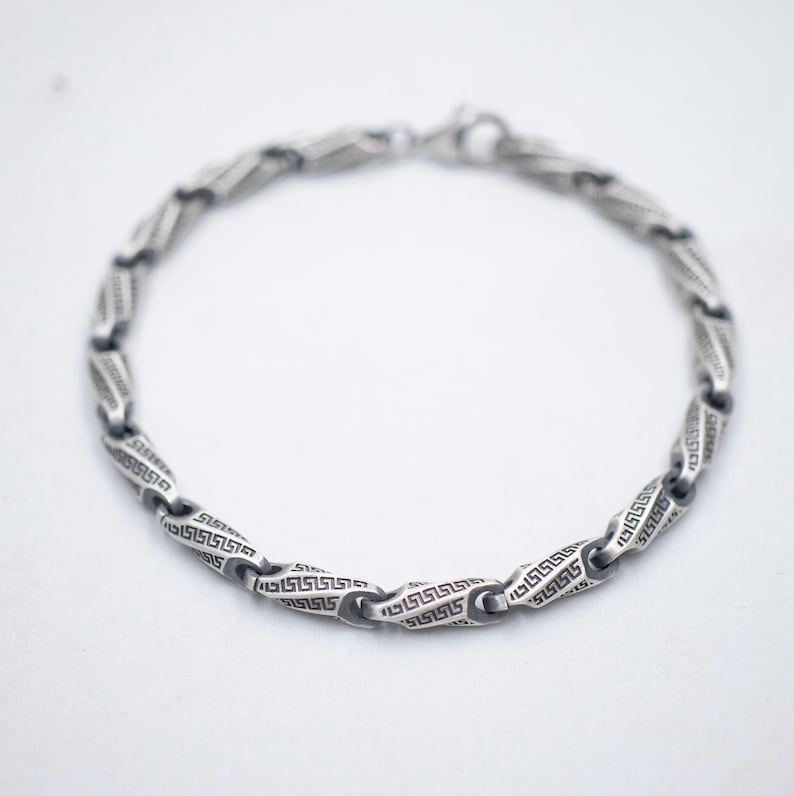 May include: A silver bracelet with a chain link design featuring a repeating Greek key pattern.