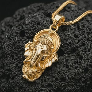 Gold plated Ganesha pendant necklace on textured volcanic rock, premium close-up studio composition emphasizing the elephant god design, crown details, and spiritual talisman symbolism.