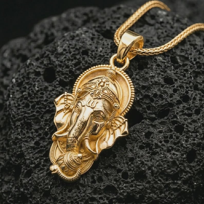 Gold plated Ganesha pendant necklace on textured volcanic rock, premium close-up studio composition emphasizing the elephant god design, crown details, and spiritual talisman symbolism.