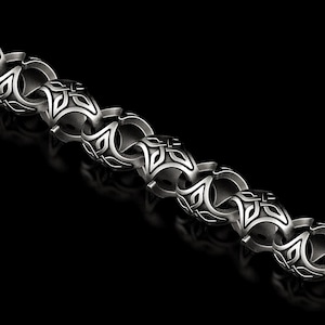 May include: A silver-coloured chain necklace featuring a repeating pattern of symmetrical designs. The links are thick and rounded, with a dark inlay. The background is black.