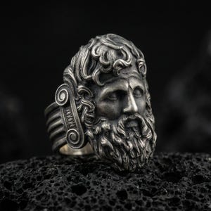 Detailed Zeus ring crafted in oxidized sterling silver, classical Greek god face with flowing beard and ornate curls, macro product shot on textured volcanic rock background