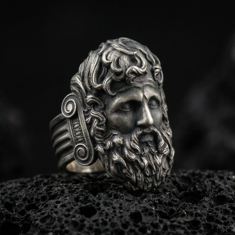Detailed Zeus ring crafted in oxidized sterling silver, classical Greek god face with flowing beard and ornate curls, macro product shot on textured volcanic rock background