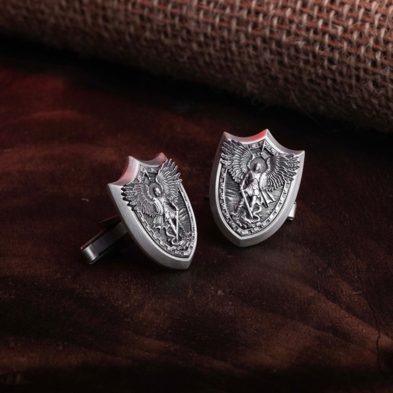 May include: A pair of silver cufflinks featuring a shield design with an embossed image of an angel with wings. The angel is holding a sword and standing on a snake.