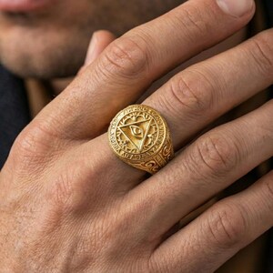 Gold plated all-seeing eye signet ring worn on a finger, close-up lifestyle image emphasizing the triangle eye emblem
