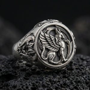 Detailed griffon ring crafted in oxidized sterling silver, winged mythical creature relief, macro product shot on textured volcanic rock background