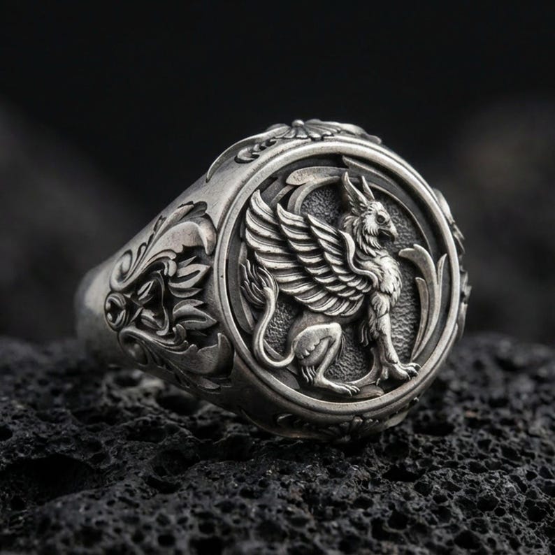 Detailed griffon ring crafted in oxidized sterling silver, winged mythical creature relief, macro product shot on textured volcanic rock background