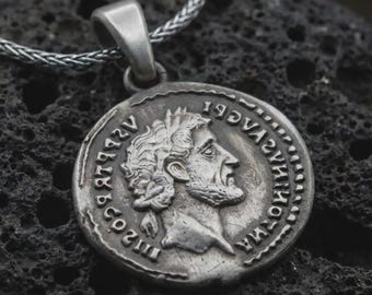 May include: A silver-colored pendant necklace featuring a detailed coin design. The coin depicts a profile of a man with a beard and laurel wreath, surrounded by ancient text. The pendant hangs from a silver chain.