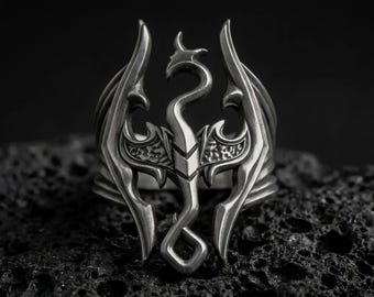 May include: A silver-toned metal ring featuring a central dragon motif with outstretched wings and a curved neck. The design includes intricate cut-out details and a textured finish on the wings, creating a bold, sculptural appearance.