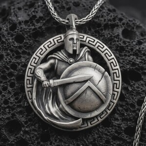 Puede incluir: Collar con colgante plateado que representa a un guerrero espartano con una espada y un escudo, rodeado por un diseño de clave griega. El colgante cuelga de una cadena plateada, mostrando detalles intrincados.