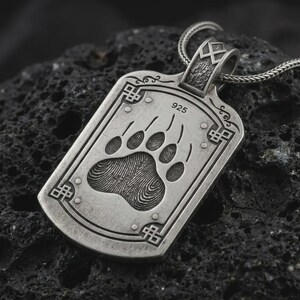 May include: A rectangular silver pendant on a chain featuring an engraved bear paw print with visible fingerprint-like texture. The pendant includes the stamp 925, decorative corner knotwork, and a textured bail with a geometric symbol at the top.