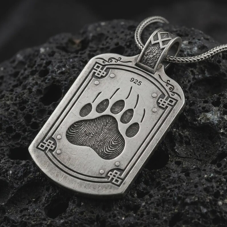 May include: A rectangular silver pendant on a chain featuring an engraved bear paw print with visible fingerprint-like texture. The pendant includes the stamp 925, decorative corner knotwork, and a textured bail with a geometric symbol at the top.