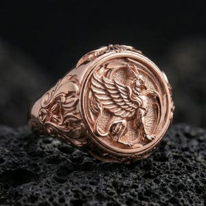 Rose gold plated griffon ring showcasing detailed winged beast engraving and refined sculptural texture, sharp close-up on volcanic rock