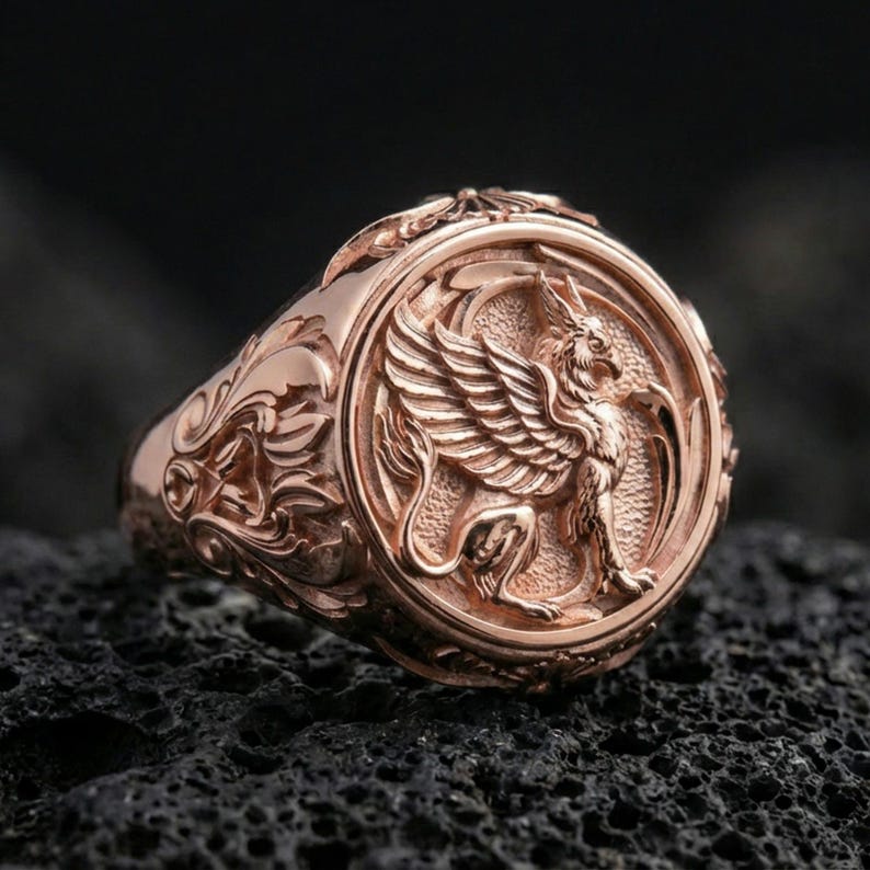 Rose gold plated griffon ring showcasing detailed winged beast engraving and refined sculptural texture, sharp close-up on volcanic rock