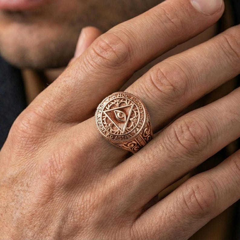 Rose gold plated eye-in-triangle ring on the finger, macro lifestyle photo showing ornate scroll engraving along the band