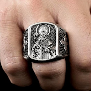 Könnte beinhalten: Silberfarbener Ring mit detailliertem Relief einer religiösen Figur, die einen Stab und ein Buch hält. Der Ring hat eine rechteckige Oberfläche mit Kreuzen an den Seiten. Die Figur trägt einen Hut und hat einen langen Bart.