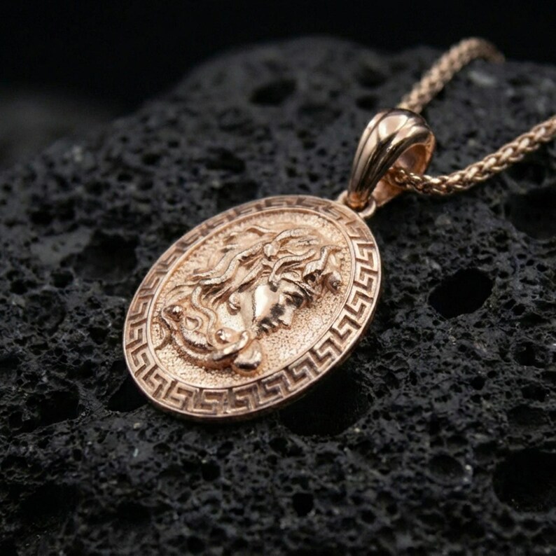 Rose gold plated Medusa necklace on dark volcanic stone, Greek key coin pendant with sculpted Medusa profile and antique-inspired finish