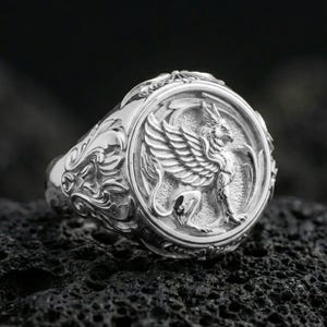 Polished silver griffon ring featuring sculpted griffin body and feathered wings, centered close-up jewelry photo on dark porous volcanic stone