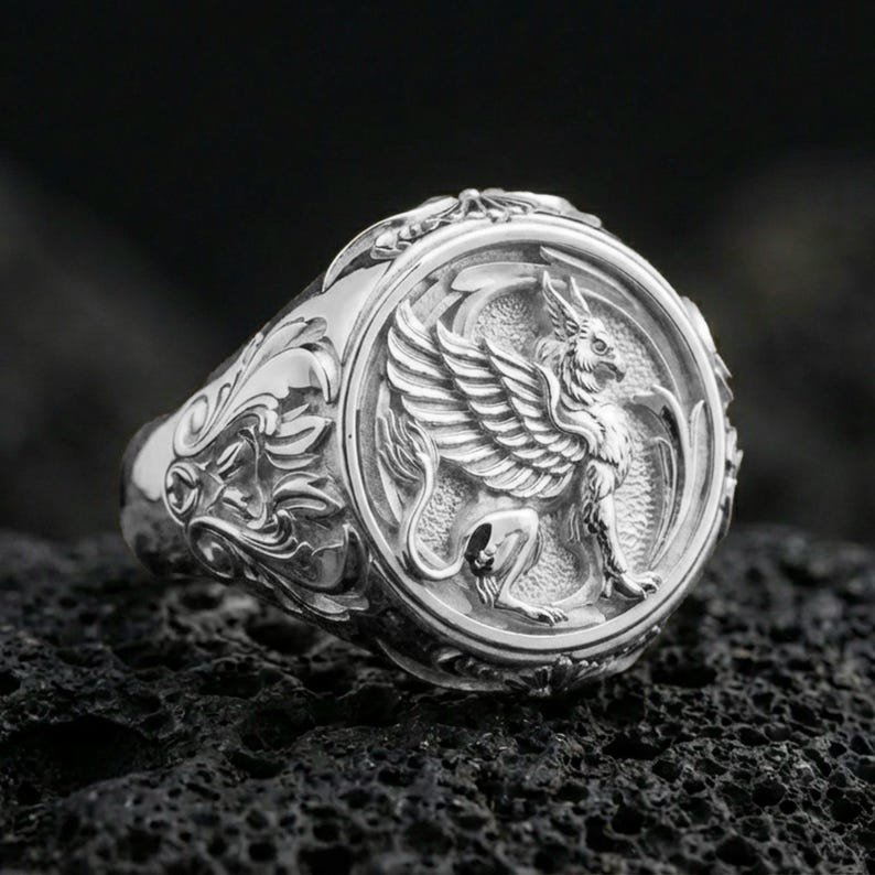 Polished silver griffon ring featuring sculpted griffin body and feathered wings, centered close-up jewelry photo on dark porous volcanic stone