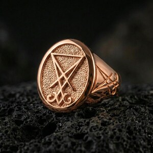 Rose gold plated Lucifer sigil signet ring with raised sigil emblem and matte-textured face, close-up on volcanic rock