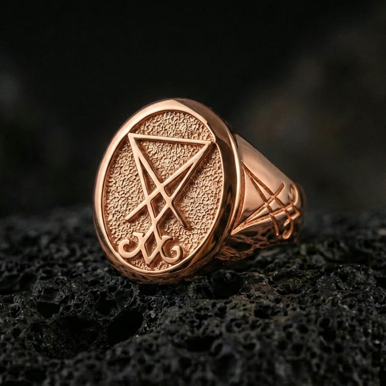 Rose gold plated Lucifer sigil signet ring with raised sigil emblem and matte-textured face, close-up on volcanic rock