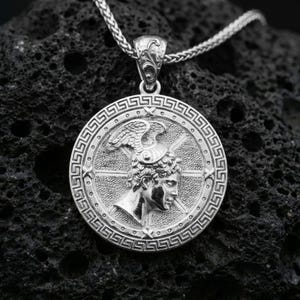Polished silver Perseus coin pendant necklace on textured volcanic rock, crisp macro product photo emphasizing clean edges, sharp engraving, and bright reflective highlights on the portrait and border.