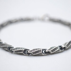 May include: A silver chain bracelet with a geometric pattern. The links are shaped like elongated ovals and are decorated with a repeating Greek key design.