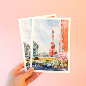 May include: Two watercolour postcards depicting a cityscape with a red and white tower in the background. The tower is surrounded by buildings and a street with cars and people. The postcards are held in a hand against a pink background.