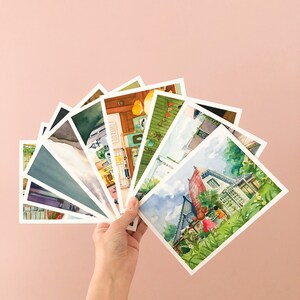May include: A hand holds a fan of watercolour postcards featuring various scenes, including a house with a red roof, a green house with a white fence, and a yellow house with a green roof.