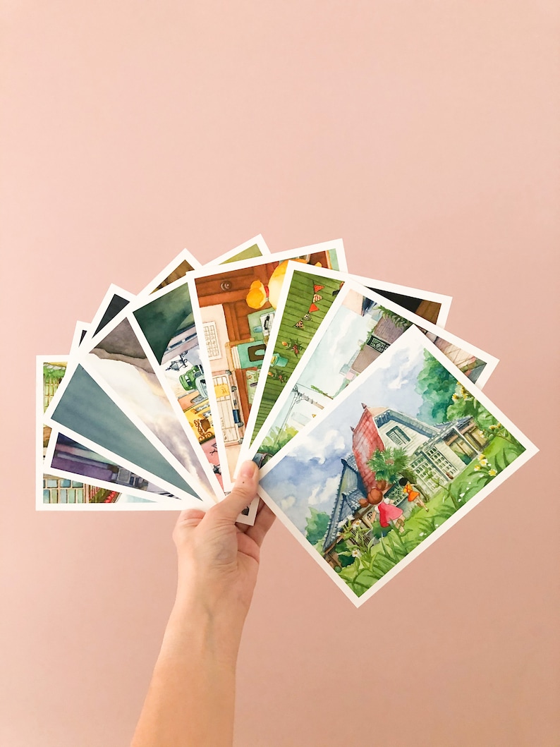 May include: A hand holds a fan of watercolour postcards featuring various scenes, including a house with a red roof, a green house with a white fence, and a yellow house with a green roof.