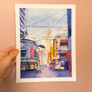 May include: A watercolour painting depicting a street scene in Japan. The painting features a tall tower, a street lined with shops and restaurants, and a large inflatable fish-shaped sign. The painting is rendered in a soft, muted colour palette.