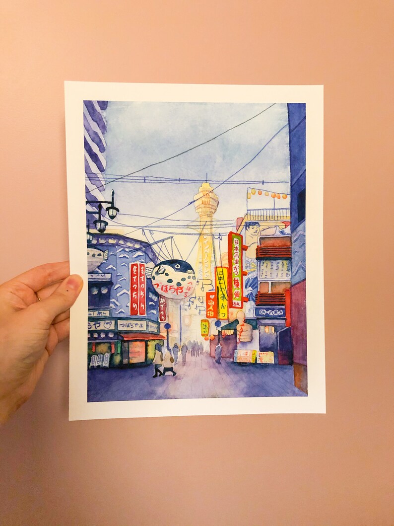 May include: A watercolour painting depicting a street scene in Japan. The painting features a tall tower, a street lined with shops and restaurants, and a large inflatable fish-shaped sign. The painting is rendered in a soft, muted colour palette.