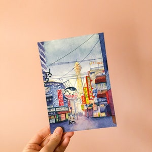 May include: Watercolour painting of a street scene in Japan. The painting features a tall tower in the distance, with buildings and streetlights lining the street in the foreground. There are people walking on the street, and the buildings have signs in Japanese.