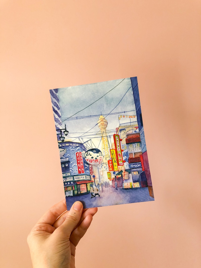 May include: Watercolour painting of a street scene in Japan. The painting features a tall tower in the distance, with buildings and streetlights lining the street in the foreground. There are people walking on the street, and the buildings have signs in Japanese.