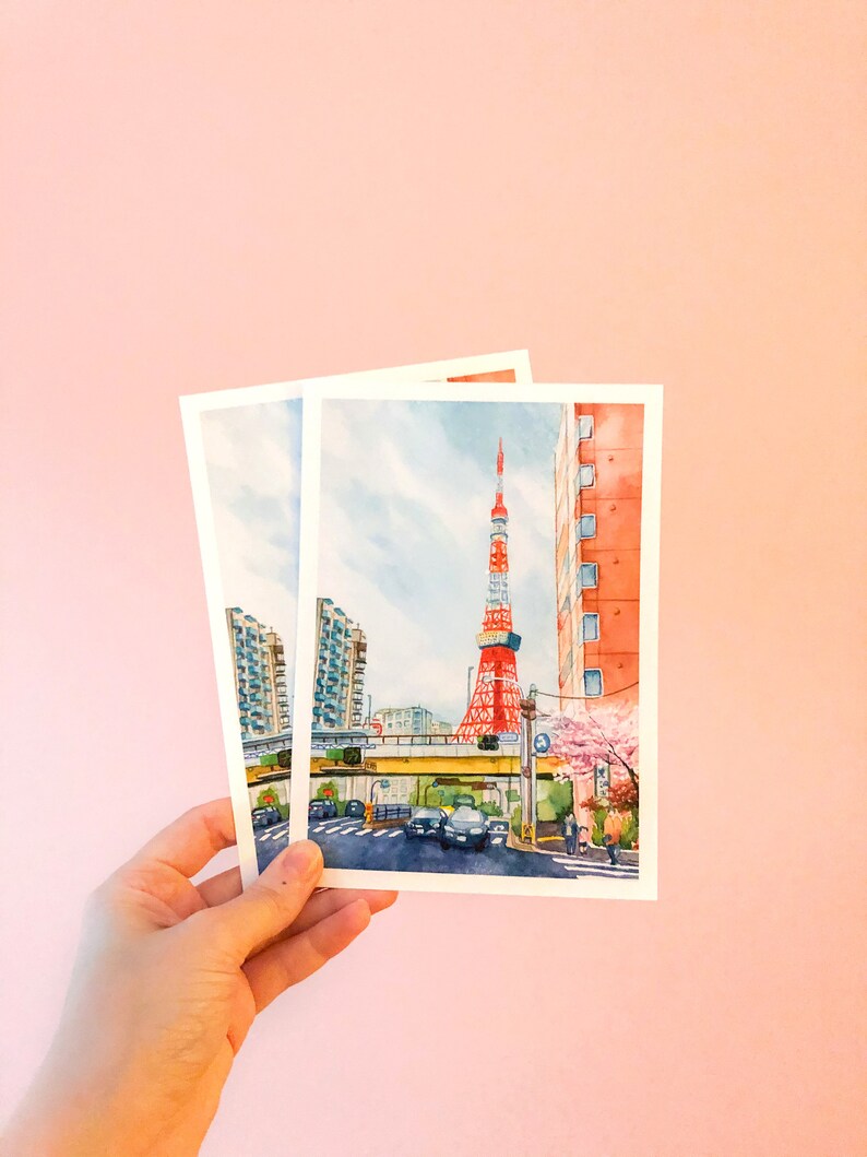 May include: Two watercolour postcards depicting a cityscape with a red and white tower in the background. The tower is surrounded by buildings and a street with cars and people. The postcards are held in a hand against a pink background.