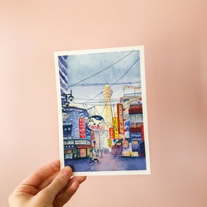May include: Watercolour painting of a street scene in Japan. The painting features a tall tower in the distance, with buildings and streetlights lining the street in the foreground. The buildings have Japanese characters on them, and there are people walking on the street.