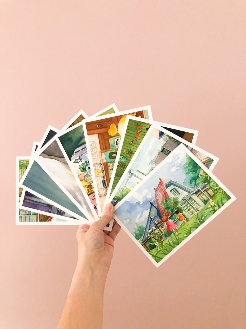 May include: A hand holds a fan of watercolour postcards featuring various scenes, including a house with a red roof, a house with a green roof, and a house with a yellow roof.