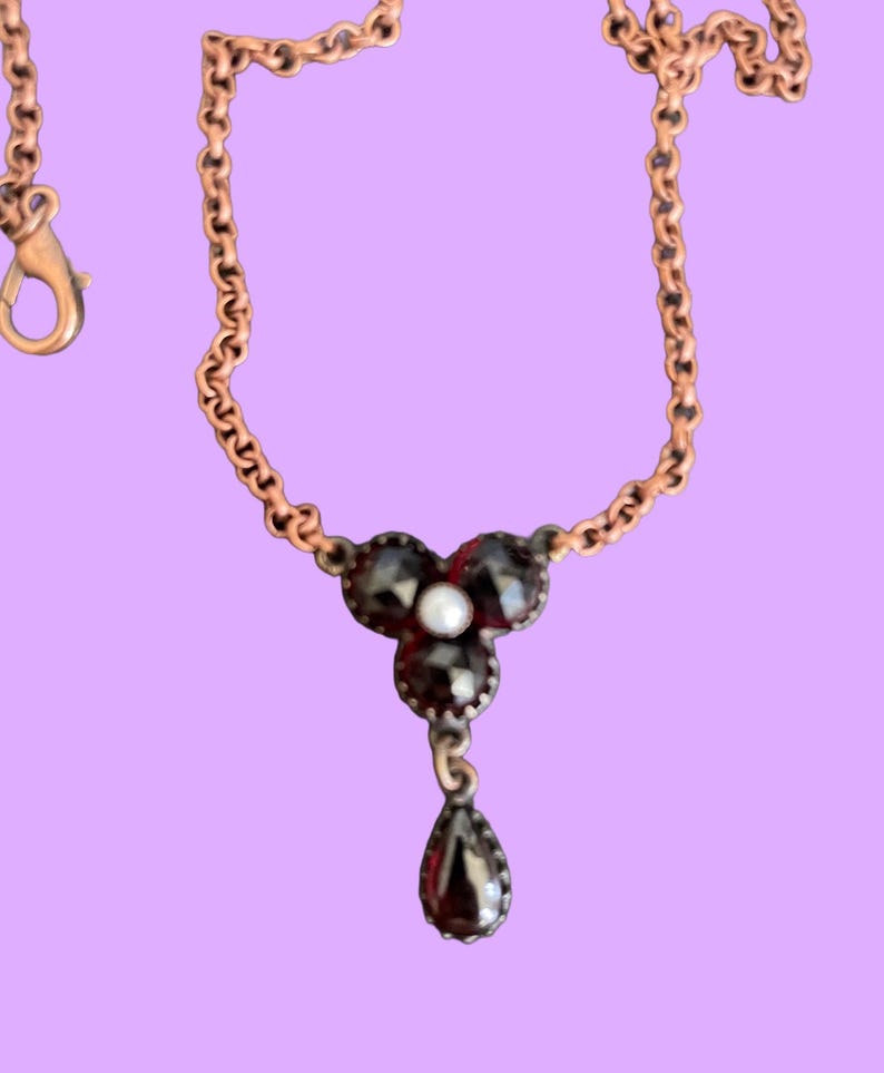 May include: A delicate necklace with a copper-colored chain and a pendant featuring three dark gemstones and a central pearl. A teardrop-shaped gemstone hangs below the pendant. The necklace is set against a lavender background.