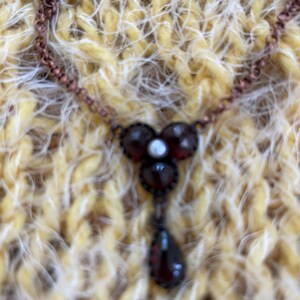 May include: A vintage-style necklace with a copper-colored chain. The pendant features three round, dark red stones with a small white stone in the center, and a teardrop-shaped stone hanging below. The necklace is displayed on a yellow and white textured surface.