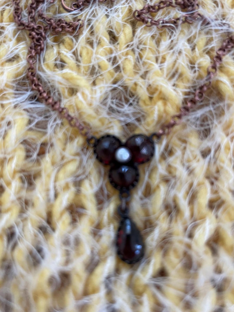 May include: A vintage-style necklace with a copper-colored chain. The pendant features three round, dark red stones with a small white stone in the center, and a teardrop-shaped stone hanging below. The necklace is displayed on a yellow and white textured surface.